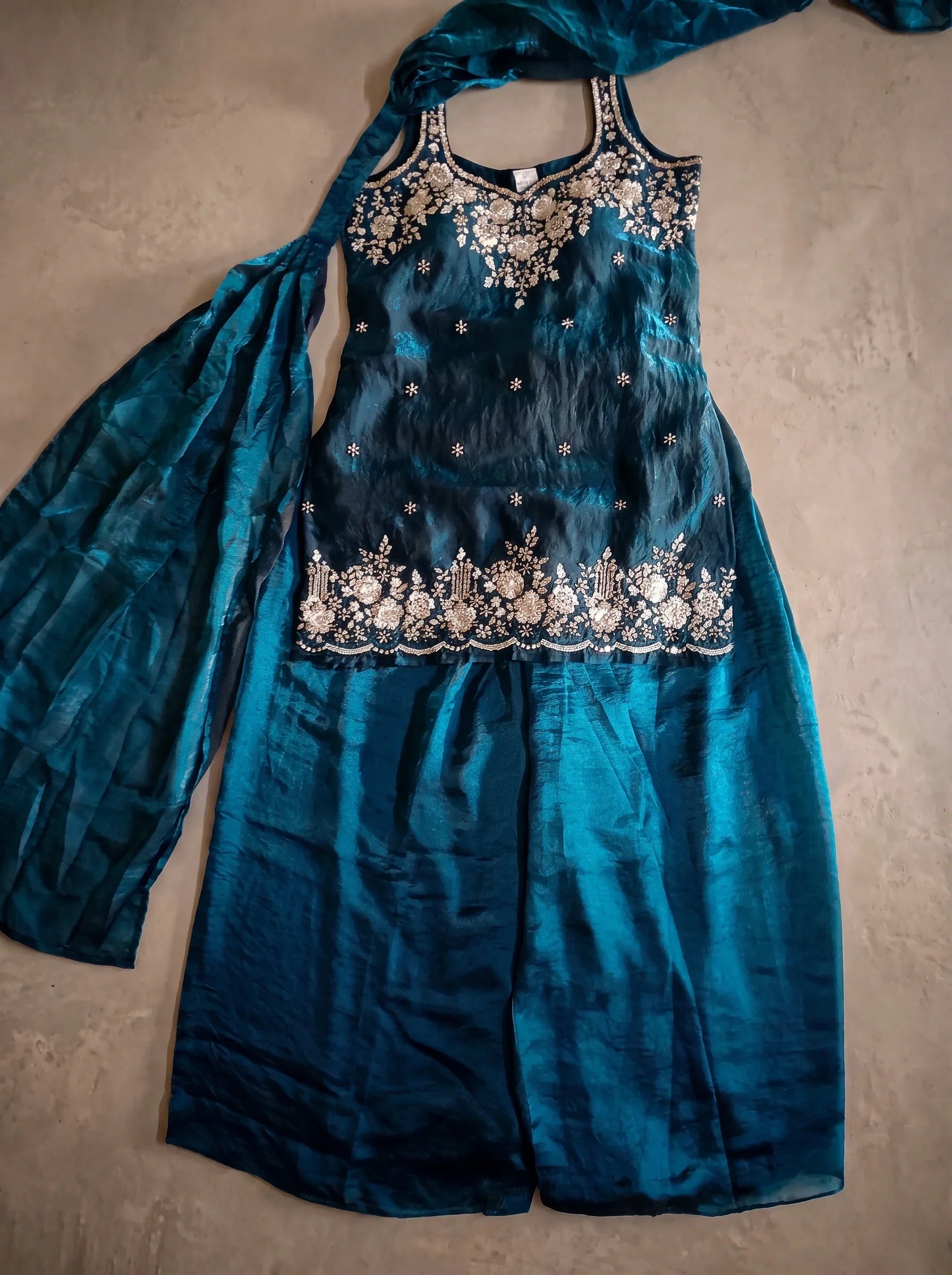 Flat lay of deep teal blue tissue silk sharara set with floral sequin and cut-bead embellished top, flared bottom and matching dupatta.