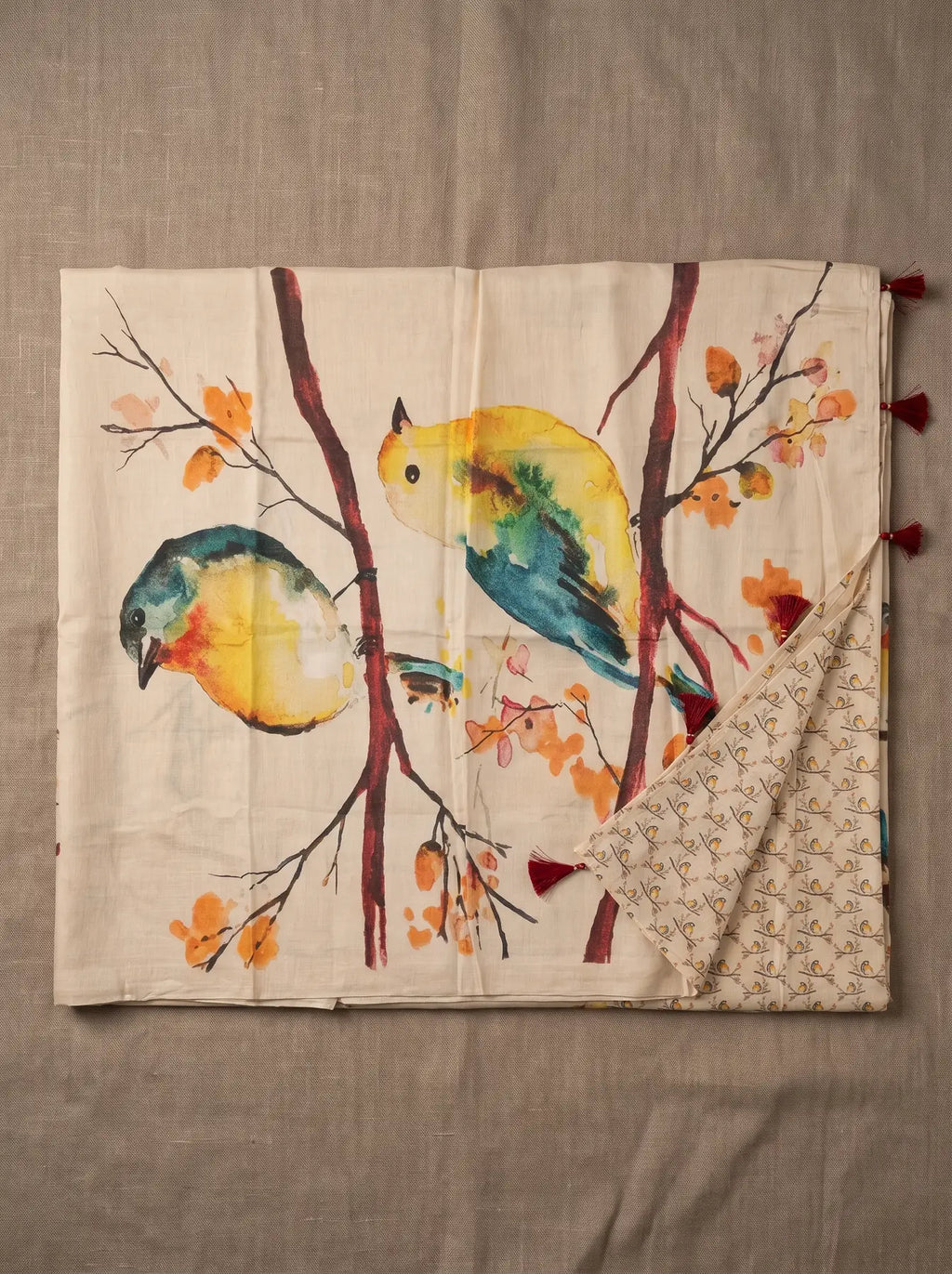 Beige linen saree displayed flat with colourful bird and branch print, printed blouse piece and maroon pallu tassels
