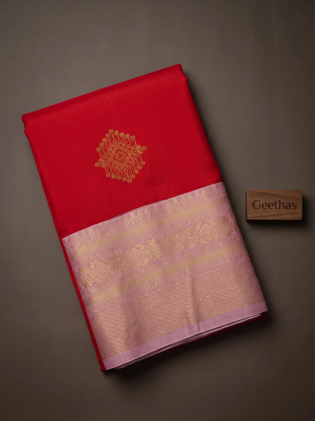 Folded red soft silk saree showing a gold-toned motif and lavender-pink woven border