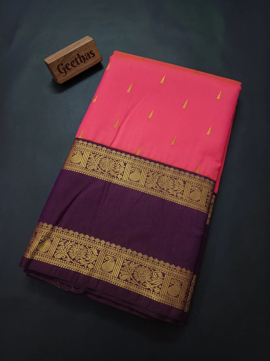Folded pink soft silk saree with deep purple border, gold-toned woven floral and geometric motifs