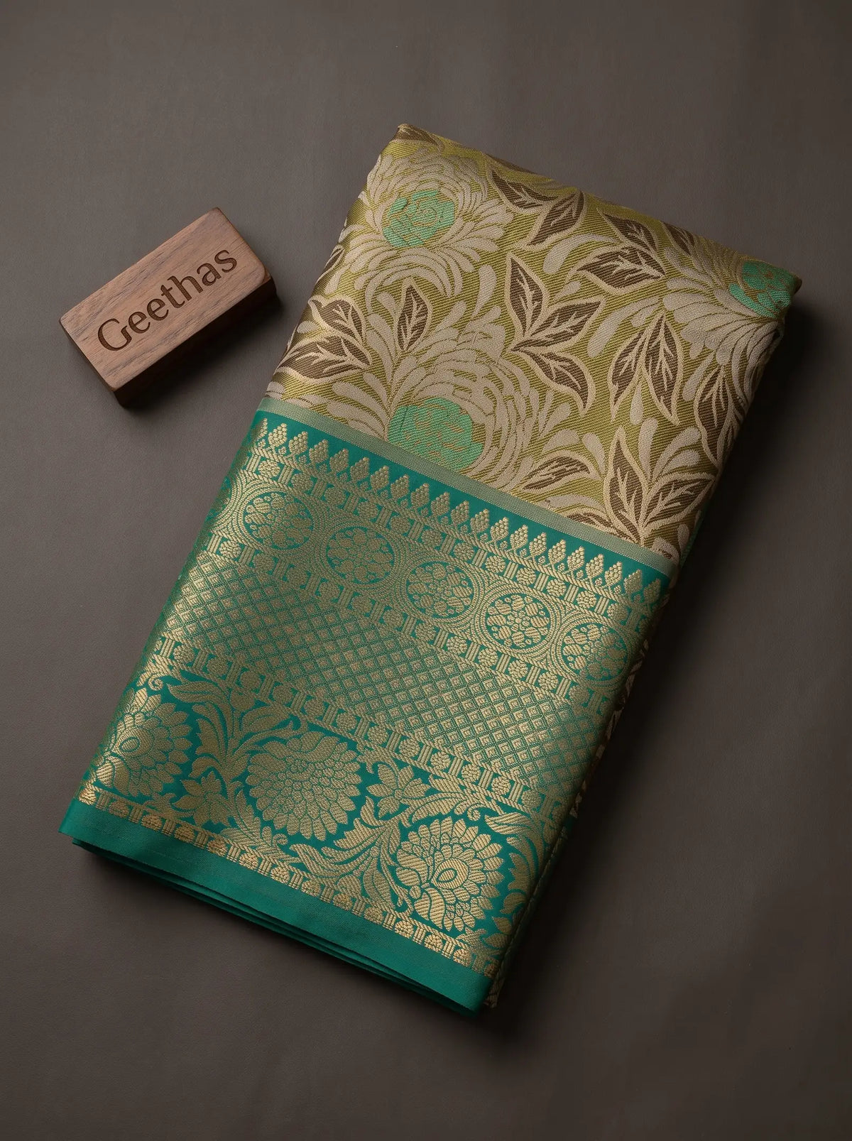 Folded beige-gold soft silk saree with floral woven motifs and a teal-green decorative border
